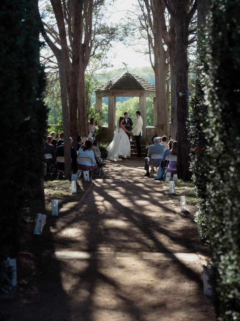 unobtrusive photographer wide angle view of wedding ceremony looking through hedges from back - documentary style wedding photography - true to life edits - natural color wedding photos