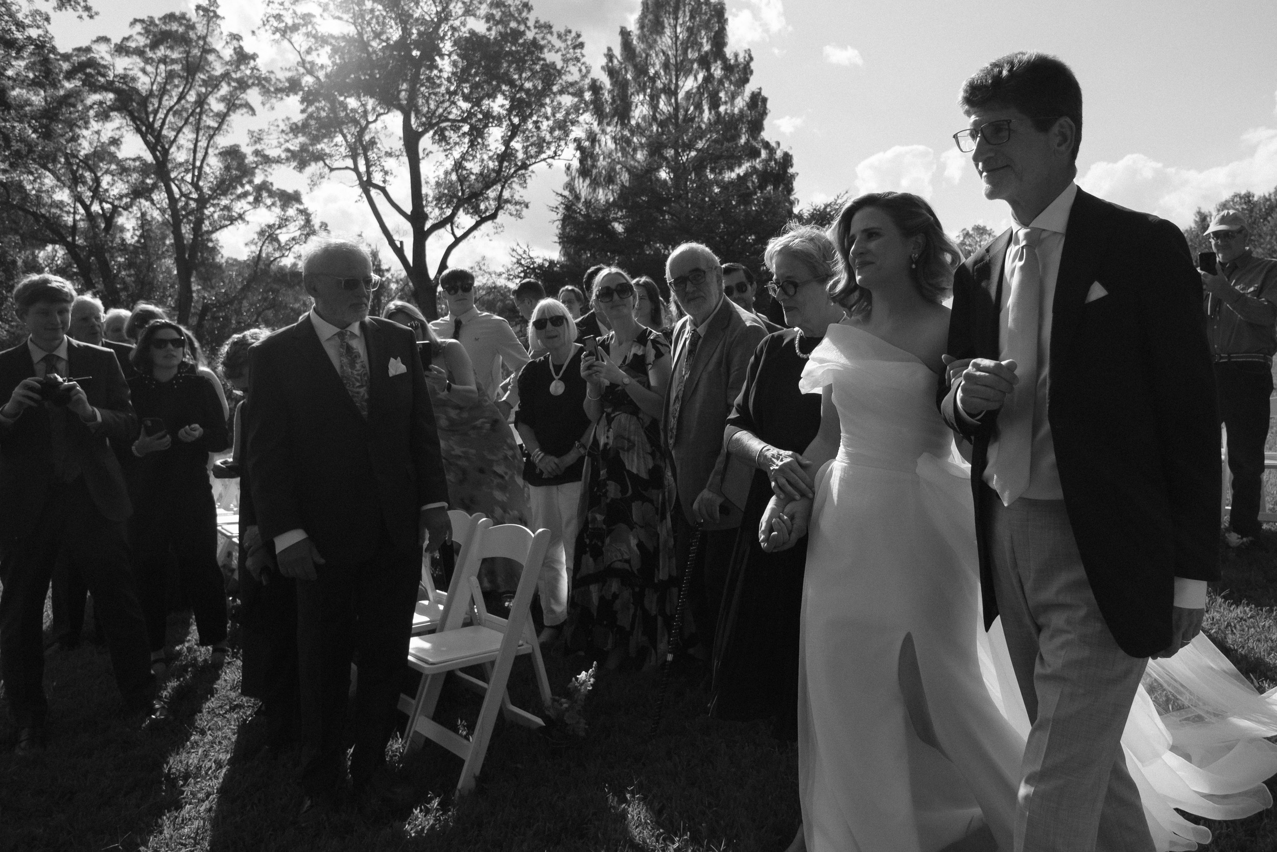 wide angle photojournalistic unstaged wedding photo bride entering ceremony with both parents - fuji acros film photography look black and white - maymont park wedding in richmond virginia - documentary photographer in richmond virginia