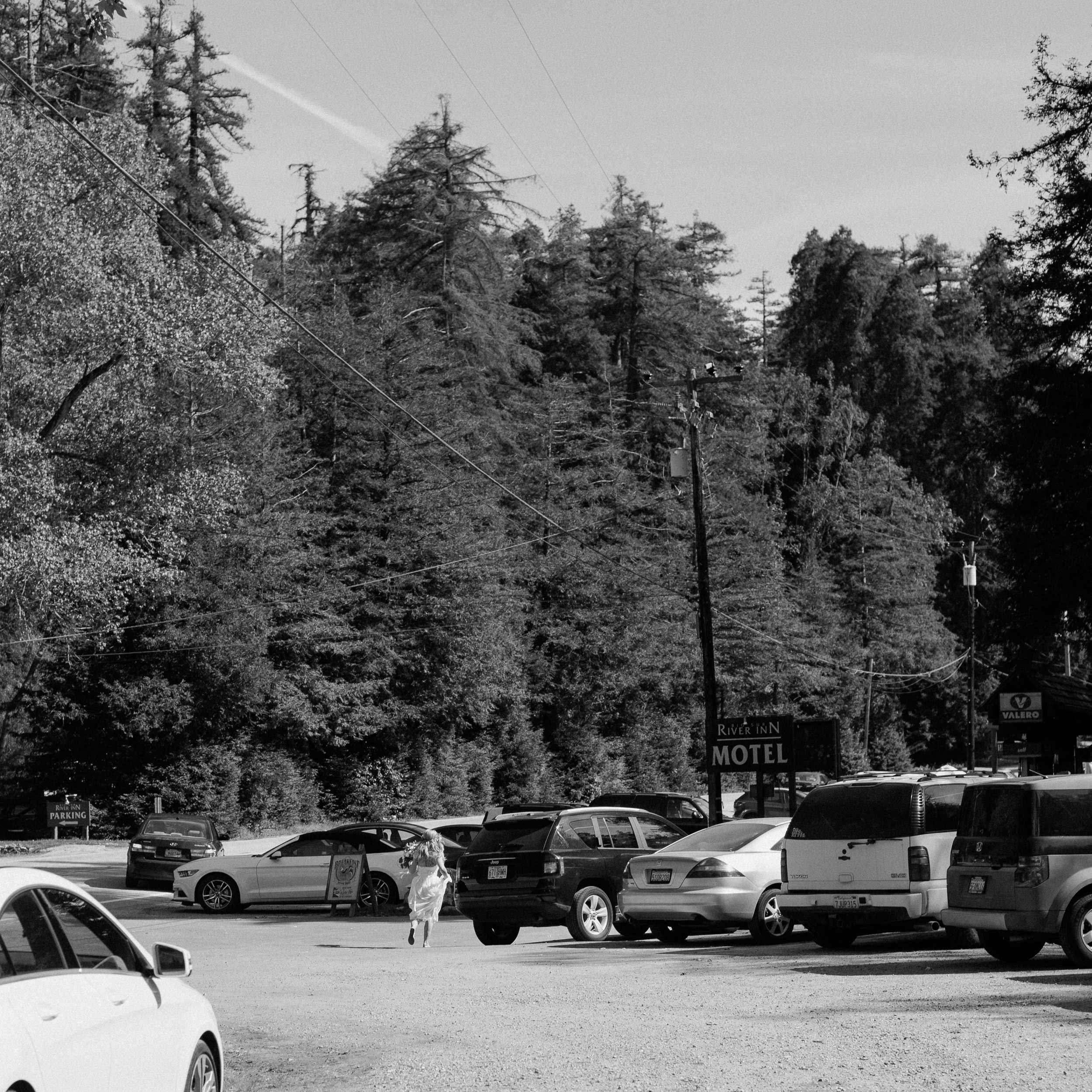 documentary style wedding photography in black and white acros film look - bride runs behind cars to hide before groom exits his room at Big Sur River Inn Motel - big sur california wedding - laid back wedding in big sur california - true to life colors