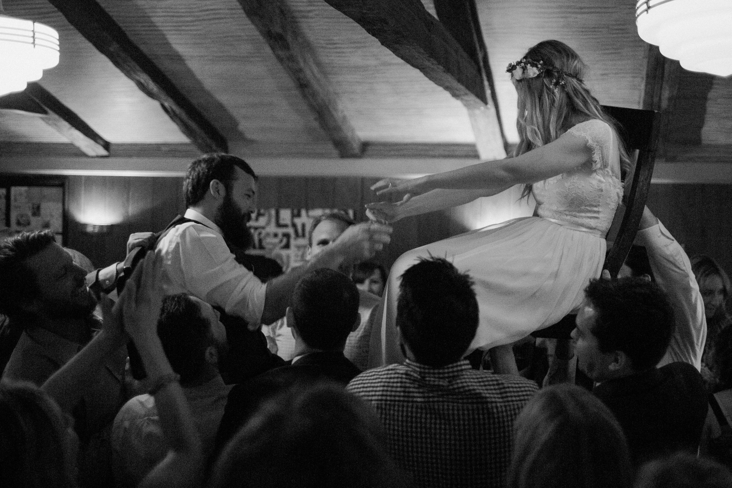 bride and groom reach for each others hands as they are lifted up in chairs during wedding reception dance at big sur bakery - documentary photographer - black and white fuji acros grainy film look